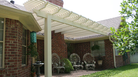 White pergola attached to a brick house over a patio area with chairs.